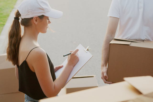 A woman removalist in a white cap and black singlet is holding a clipboard and ticking off an item with a black pen. The ground around her is piled high with cardboard boxes and a man in a white shirt is carrying a box in front of her. Image at LondonOfficeSpace.com.