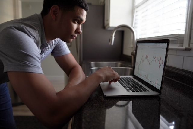 A man in a grey t-shirt checks a graph of market conditions on his laptop in the kitchen. His laptop is resting on the kitchen bench next to the sink and he's standing, leaning on the bench. Image at LondonOfficeSpace.com.