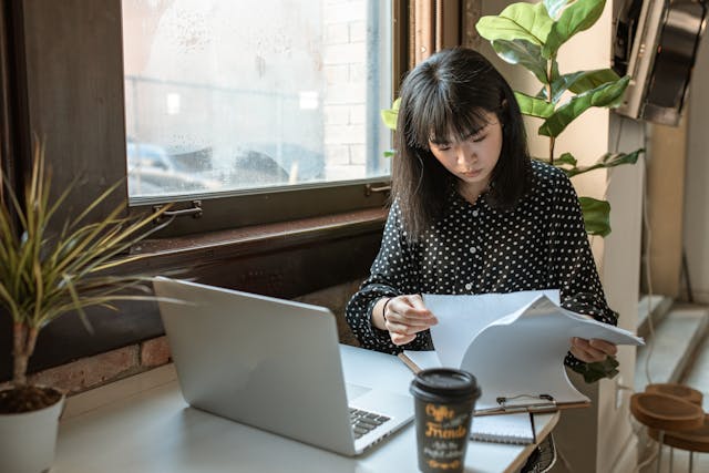 A woman with black hair is sitting at a desk and looking through documents on a clipboard. Her silver laptop and takeaway coffee cup sit on the desk in front of her. Image at LondonOfficeSpace.com.