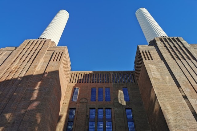 A ground-up view of Battersea Power Station's art deco facade and two of its disused white chimneys that rise into a blue, cloudless sky. Image at LondonOfficeSpace.com.