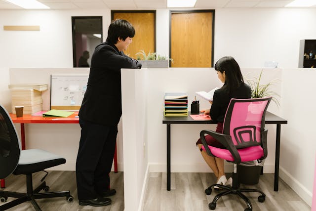 A man in a black business suit is resting his arms on his cubicle's partition and talking to his female colleague, who is sitting at her desk in the neighbouring cubicle. Image at LondonOfficeSpace.com.