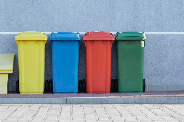 A yellow, blue, red, and green recycling bin are lined up on a narrow footpath in front of a light, brick paved street. Image at LondonOfficeSpace.com.