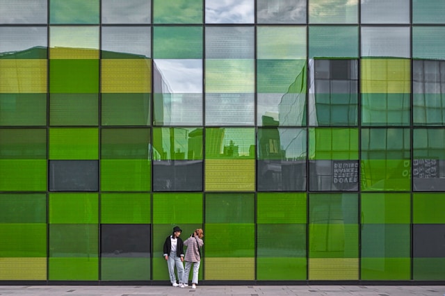 A woman in casual wear and a black bucket hat leans casually against a glass faced office building composed of rectangles in many shades of green. Her friend is taking a photo of her with her phone. Image at LondonOfficeSpace.com.