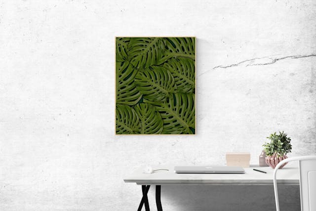 Artwork of interlocking leaves placed on a rustic white wall. Below is a white chair and desk with a silver closed laptop and small potted plant resting on it. Image at LondonOfficeSpace.com.