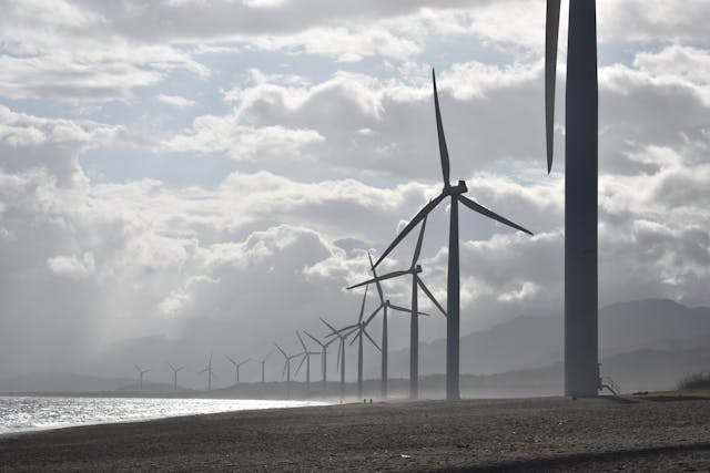 A daytime, sea-level view of a long stretch of coastline with tall wind turbines lining it. Mountains are just visible in the background, and sunlight glints off clouds that seem to almost fill the sky. Image at LondonOfficeSpace.com.