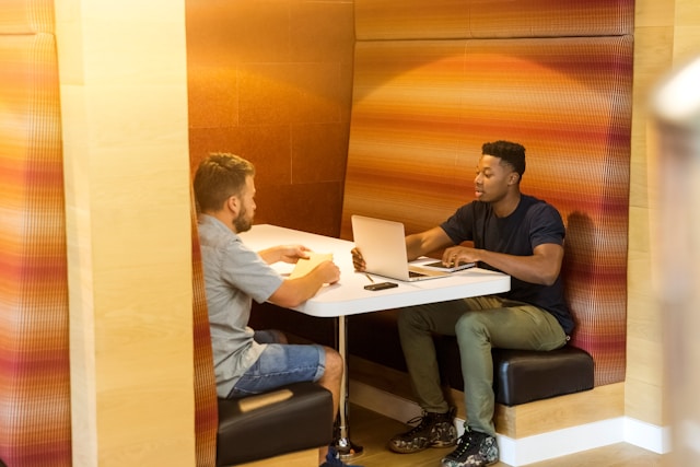 Two businessmen in smart casual dress work across from each other in a modular privacy booth. One in a black T-shirt and green trousers works on a silver laptop, and his colleague in a grey, short-sleeved collared shirt holds a notepad. Image at LondonOfficeSpace.com.