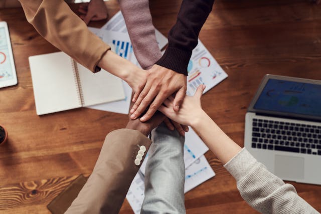 A team of six people place their hands on top of each other across a desk in a team hand-stack. The wooden desk is littered with a laptop, tablet, phone, coffee mug, and various papers and notepads. Image at LondonOfficeSpace.com.