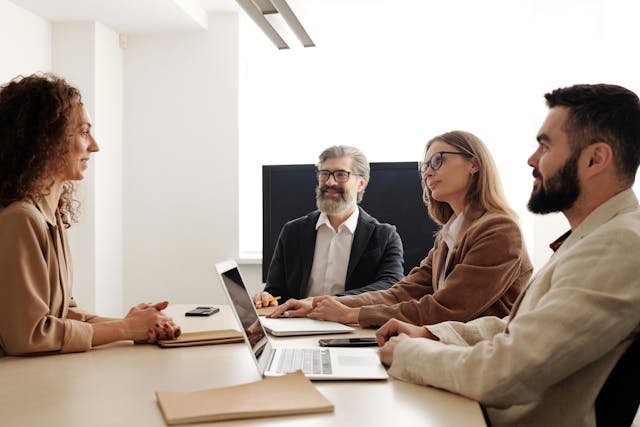 A businesswoman with curly hair is seated at a meeting room table being consulted by three office space consultants, comprising two men and one woman, about what she needs in an office space upgrade. Image at LondonOfficeSpace.com.