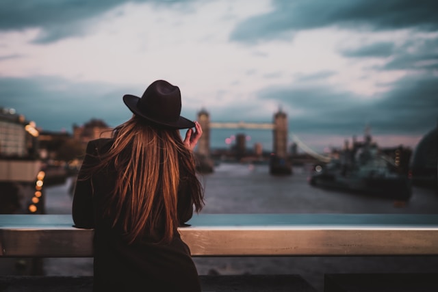 A stylish businesswoman with red hair in a black jacket and wearing a black, rimmed hat stands on a bridge over the River Thames at dusk, leaning on the railing and looking towards London’s Tower Bridge. Image at LondonOfficeSpace.com.