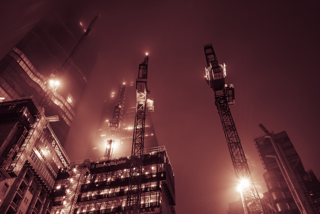 A nighttime view from the ground up towards commercial building construction occurring at Bishopsgate in Central London. Lights from midpoints on the three to four visible cranes light the scene, as well as lights from some levels within the buildings being constructed. The sky is shrouded in either clouds or fog, which casts a reddish hue to the view. Image at LondonOfficeSpace.com.