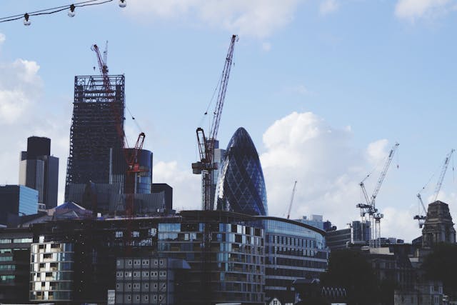 A daytime, close-up view of the central London skyline with The Gherkin and The Cheesegrater, or, more officially, the Leadenhall Building, featuring prominently under a cloudy, light blue sky. Many building cranes also poke up from the city skyline towards the sky. Image at LondonOfficeSpace.com.