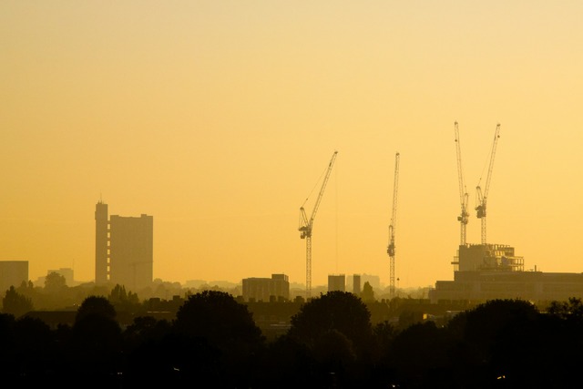 A dusk view towards West London’s Trellick Tower, which is in the distance on the left, and there are four building cranes poking up towards the hazy orange sky, surpassing the tower’s height on the right. Image at LondonOfficeSpace.com.