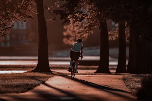 A man wearing a t-shirt, shorts, and dark helmet rides a bike along a path within a park at dusk. Autumnal trees with orange and red leaves line the path, and the setting sun casts long shadows from the trees across the path. Image at LondonOfficeSpace.com.