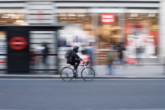 An office worker wearing warm, dark clothes, a grey backpack, and a black helmet rides a white road bike along the side of the road past a London bus stop shelter and a glass-fronted shopfront. Image at LondonOfficeSpace.com.