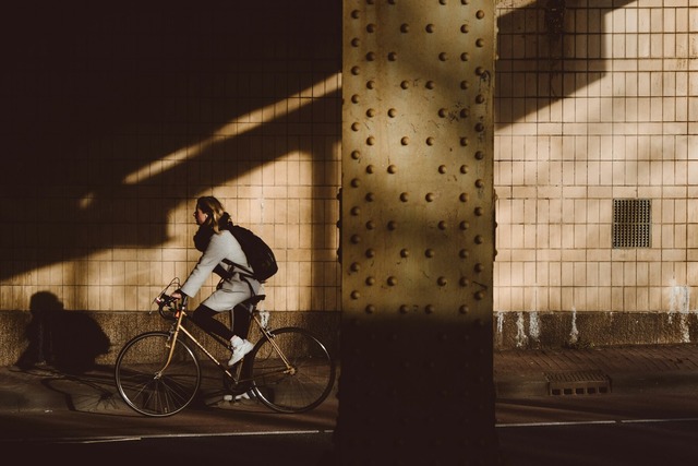 A blonde woman wearing a light grey jacket, black scarf, and black backpack rides a retro gold chrome coloured bicycle along a bike path that runs under an overpass. Image at LondonOfficeSpace.com.