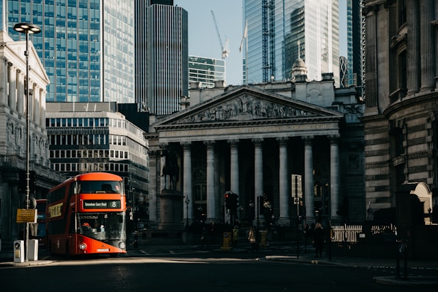 A daytime view from the street towards the shadowed, old, and columned Royal Exchange building in London’s financial district. One of London’s iconic red double-decker buses pulls out from a bus stop to the left of the Royal Exchange and emerges into sunlight cast from between tall buildings all around. Many shiny glass-faced skyscrapers rise up in the background behind the Royal Exchange. Image at LondonOfficeSpace.com.