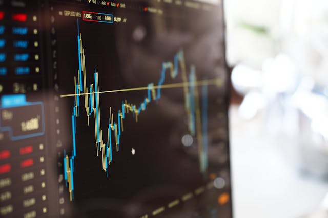 A close-up view of an undulating graph of the stock market on a computer monitor. The graph is displayed in dark mode, so the lines of the graph are bright blue and yellow on a black background. Image at LondonOfficeSpace.com.