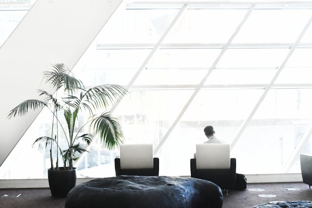 A businessman in a white collared shirt sits by himself on a modern black-and-white chair, looking towards long, diagonal windows that let bright sunlight stream in high up in an office building. There is another matching empty chair to his left, as well as a tall indoor plant in a black pot. Image at LondonOfficeSpace.com.