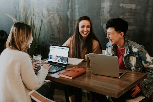 Three businesswomen in casual dress are laughing while collaborating and sitting at a shared desk with their laptops, drinks, and note-taking implements in front of them. Image at LondonOfficeSpace.com.