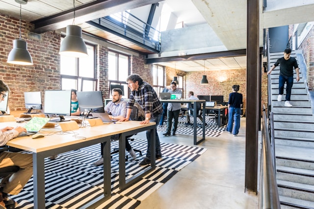 A view down the length of a modern, well-lit and open-plan workspace with exposed brick walls, polished concrete floor, steel beams, and a high vaulted ceiling. Many people are working at shared desks, either standing or sitting in front of their laptops or computers. Image at LondonOfficeSpace.com.