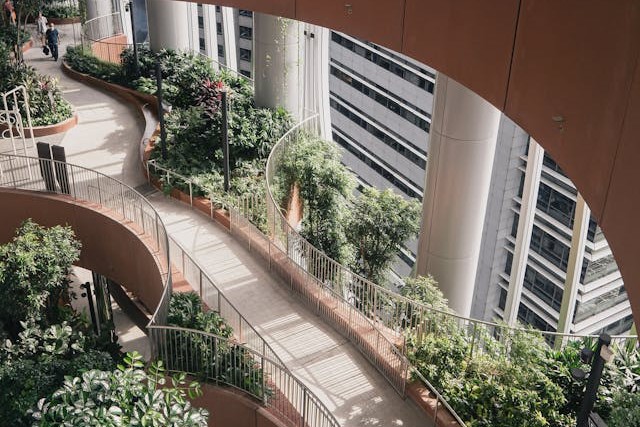 A view down on a raised, winding path connecting different parts of a modern office tower. A few businesspeople are walking down the path at the top of the image, and the path is lined with abundant plants and trees that rise up from the lower levels of the building. Image at LondonOfficeSpace.com.