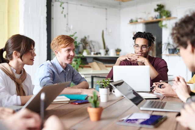 A team of six Generation Z employees in smart casual attire sit around a large wooden desk having a meeting. Some of them have their laptops in front of them, and others have note-taking implements. The room they are in is well-lit with natural light, and many pot plants adorn shelves along the whitewashed walls. Image at LondonOfficeSpace.com.