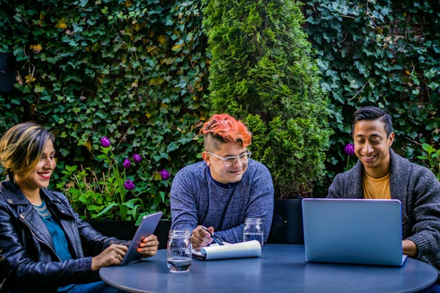 Three Gen Z workers in smart casual attire sit outside at a round table with a backdrop of lush green plants covering the wall and there are some purple flowers growing up from the base of the wall. The woman on the left is smiling and wears a leather jacket and has a tablet in her hands, the smiling man in the centre is holding a pen with his hand resting on an open notebook and has a trendy dyed red hairstyle with the sides of his head shaved, and the man on the right is smiling and working on a laptop. Image at LondonOfficeSpace.com.