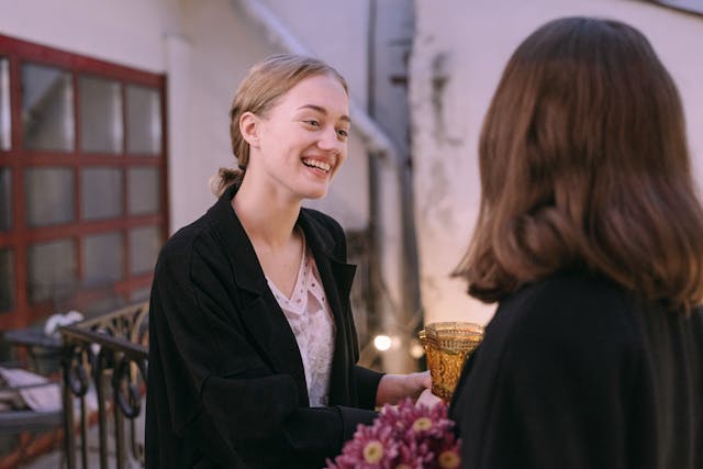 Two young businesswomen in dark blazers are standing and conversing. One of the women with her back facing the viewer has dark brown hair hanging down to her shoulders, and the blonde woman facing her is smiling broadly and holding a glass with clear liquid in it. Image at LondonOfficeSpace.com.