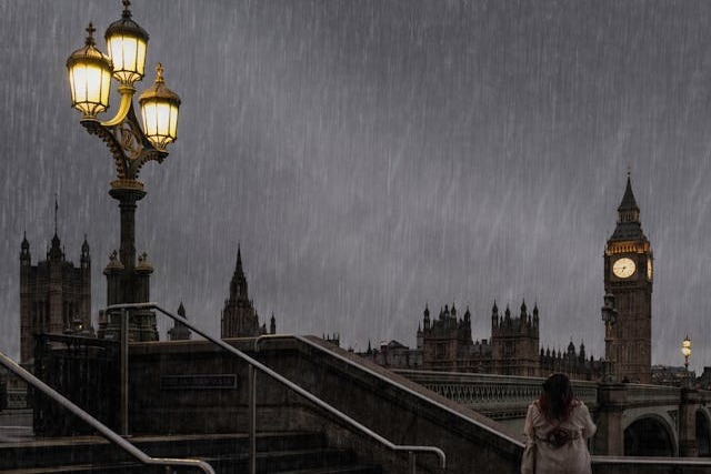 A nighttime view of a woman standing at the end of a bridge looking towards Big Ben and the Gothic spires of Westminster Hall and the House of Lords across the river. The sky is dappled grey and sheets of rain are pouring down. The primary light sources are Big Ben’s lit clock face and the antiquated light post with three lantern-like lights lit to the woman’s left. Image at LondonOfficeSpace.com.