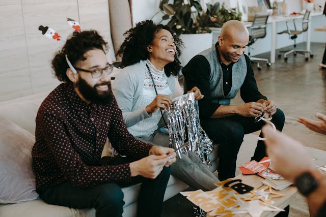 Three colleagues, two men and a woman between them, in smart casual dress, sit on a couch in the office smiling as they create Christmas decorations out of various materials that are on the coffee table in front of them. Image at LondonOfficeSpace.com.