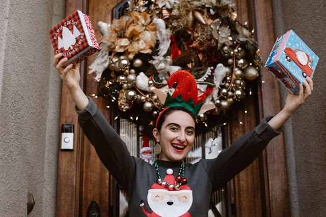 A joyous woman with dark hair wearing a Christmas elf hat, a green jumper with an image of Santa on the front and a necklace with shiny gold and red baubles on it is standing on the doorstep with a house’s front door behind her. She’s holding up two gifts, one in each hand, in the air and smiling widely. The front door has an elaborately decorated Christmas wreath hung on it. Image at LondonOfficeSpace.com.