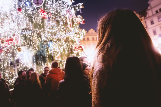 A woman in a warm knitted top looking toward a tall and wide Christmas tree in the middle of a city square at night. The tree is covered with white fairy lights, and gold and red baubles are scattered around the branches. Groups of people mill about between the woman and the tree. Image at LondonOfficeSpace.com.