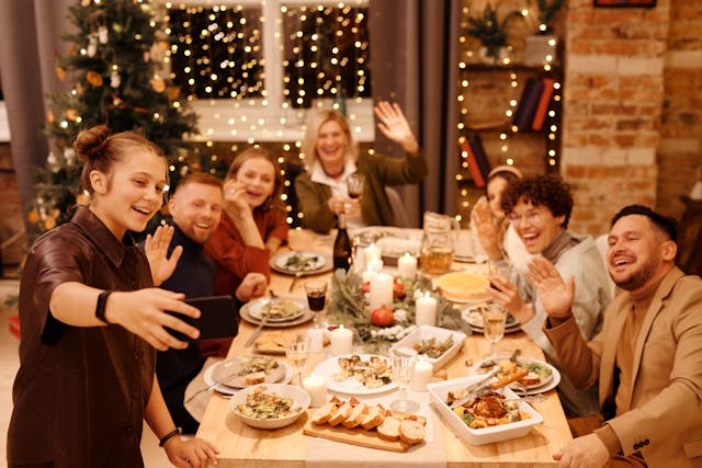 A group of seven colleagues in smart casual clothing sit around a wooden dining table that’s heavily laden with an assortment of foods and beverages, and Christmas decorations surround them, including a Christmas tree in the corner of the room and fairy lights hanging from the window behind them. One of them, a woman in a short-sleeved, black collared shirt, stands at the head of the table holding out her phone’s camera so that their remote colleagues can join in the festivities. Everyone around the table is smiling, and most are waving to the phone’s screen. Image at LondonOfficeSpace.com.