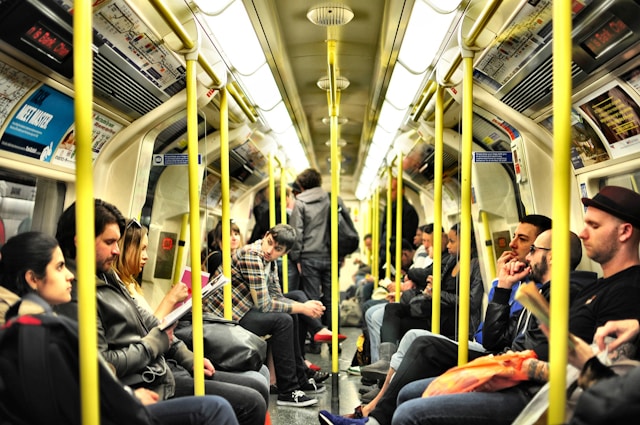 A view down the centre aisle of the carriage of one of the London Underground’s trains. The interior is packed with commuters, all the seats on either side are full, and some people are standing in the centre aisle and near the doors for entry and exit. Most of the people are wearing long clothing and some are reading books, holding bags, or looking at their phones. Image at LondonOfficeSpace.com.