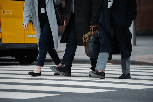 A close-up view of the bottom halves of three smartly dressed colleagues walking across the white lines of a pedestrian street cross-walk to go to work in the morning. The two men on the right carry soft leather briefcases and wear long black coats, and the woman on the left has a large shoulder bag and wears a long grey coat. The woman on the left and the man on the far right of the three have lanyards with their work IDs visible hanging down from their necks, and the rear of a yellow car is visible parked on the side of the street behind the woman and to the left. Image at LondonOfficeSpace.com.