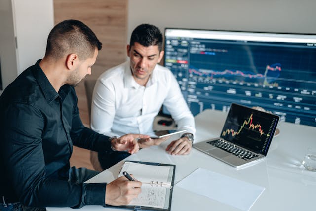 An image of two male office space brokers sitting next to each other at a white desk discussing market dynamics and how to secure the best deal for a client. One in a dark collared shirt is writing in a notebook and gesturing with his free hand toward the chrome tablet that the other man in a white shirt is holding. An open laptop is on the desk showing the peaks and troughs of a market’s line graph, and there’s a big screen on the wall behind them showing another graph and market data. Image at LondonOfficeSpace.com.