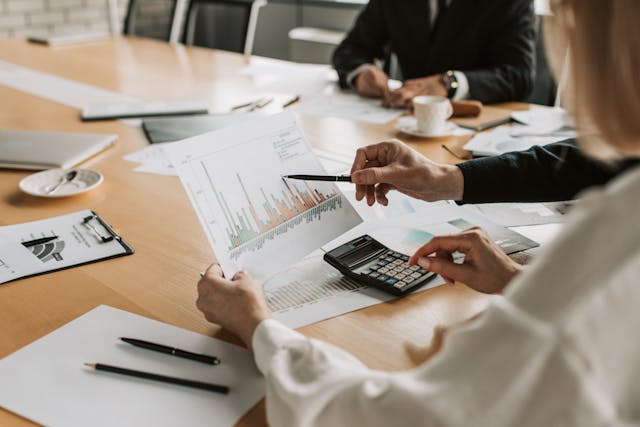 Business leaders sit around a large, light wooden desk in a boardroom, going over their operational data to determine their office space requirements. The businesswoman in the foreground is holding up a printed bar graph with her left hand and entering figures into a calculator with her right hand, while her colleague to her right is pointing at a data point on the graph with a black pen. Image at LondonOfficeSpace.com.