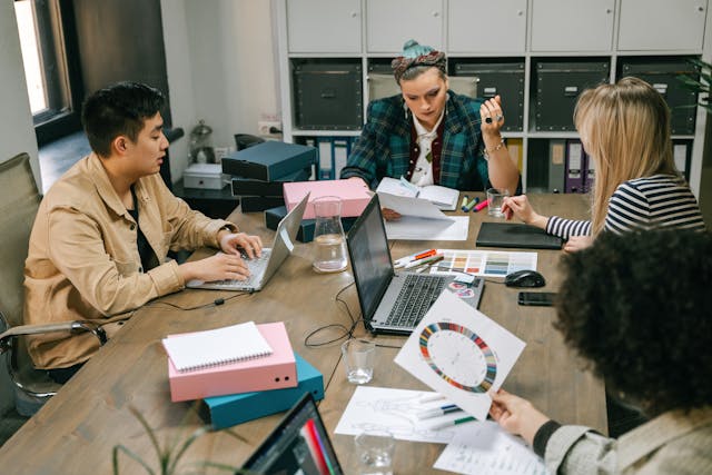 Colleagues in a creative business sit around a wooden meeting room table discussing their office space requirements. Many colour swatches, files, coloured pens, and their laptops are on the desk. Image at LondonOfficeSpace.com.