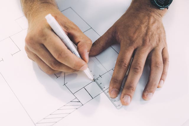 A close-up view of a man’s hands marking out an office space floor plan on white paper with a black pen and clear plastic ruler, after determining the optimal office space layout for his business. Image at LondonOfficeSpace.com.