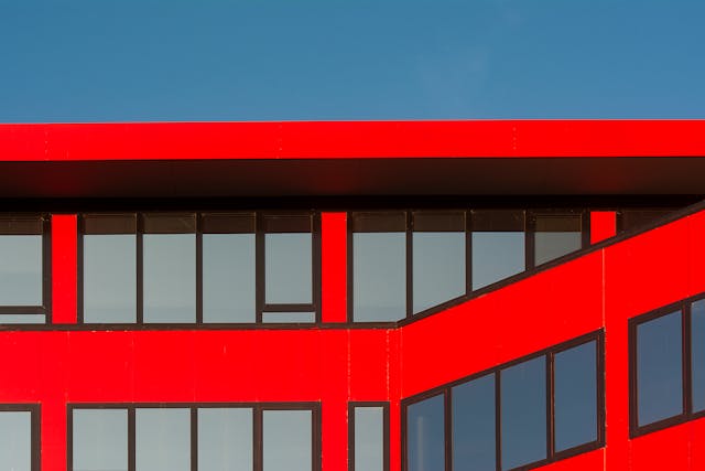 A close-up view of the top couple of floors of an office building with a red facade. The many rectangular windows have a matte greyish-blue shine, and a strip of blue, daytime sky is visible above the top of the building. Image at LondonOfficeSpace.com.