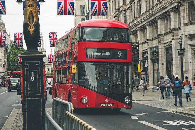 A front-on view of one of London’s iconic red double-decker buses driving up a street in the Hammersmith area of London. People are walking down the footpaths on the sides of the road, which are lined with historical buildings, and decorative Union Jack flag bunting hangs across the street from the buildings on either side of the street. As an area on the outskirts of London, the cost of office space in Hammersmith is more affordable for many startups’ survival. Image at LondonOfficeSpace.com.