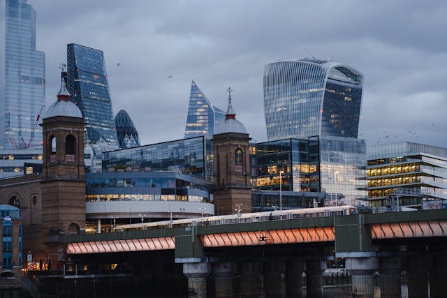 An early evening view from next to a bridge towards London’s city core, with a few of the city’s famous skyscrapers – including The Cheesegrater, Walkie-Talkie and Gherkin buildings – peeking up above the buildings closer to the bridge. An overland train is going across the bridge, some lights are coming on in the office buildings to cut through the dusk gloom, and a few birds are flying through the cloud-filled sky. The cost of office space in London’s centre is relatively high, even by global standards, so if startups hope to survive renting office space here, they tend to require substantial injections of capital and to be in the finance, insurance, or legal fields to make the most of proximity to key clients and supporting businesses. Image at LondonOfficeSpace.com.