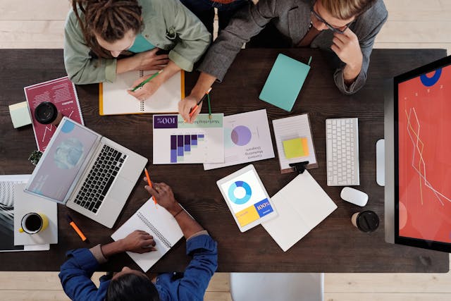 A top-down view of three startup colleagues having a meeting about their financials to ensure the young business's survival. They are sitting around a large rectangular wooden meeting table. The table is littered with printed graphs and their note-taking materials, and there is a computer monitor, keyboard, and mouse at the right end of the desk showing a red graph with white lines on it trending upwards. The woman at the bottom of the image is holding a red pencil, resting her hands on her open notebook, and has an open laptop and mug of black coffee in front of her. The woman sitting across from her is holding a green pencil poised to write in her open notebook, while her male colleague points to a printed bar graph in the middle of the table. Image at LondonOfficeSpace.com.