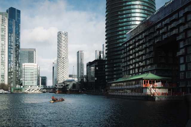 A small passenger boat for tourists, with its red-garbed pilot standing at the stern, is guided down one of the calm waterways in the Canary Wharf area of London. Skyscrapers of different sizes and designs rise from the banks, and their windows shimmer under a blue, daytime sky mottled with some greyish-white clouds. Canary Wharf and many other London areas in its vicinity are more budget-friendly when seeking out offices to rent compared to more central London locations. Image at LondonOfficeSpace.com.