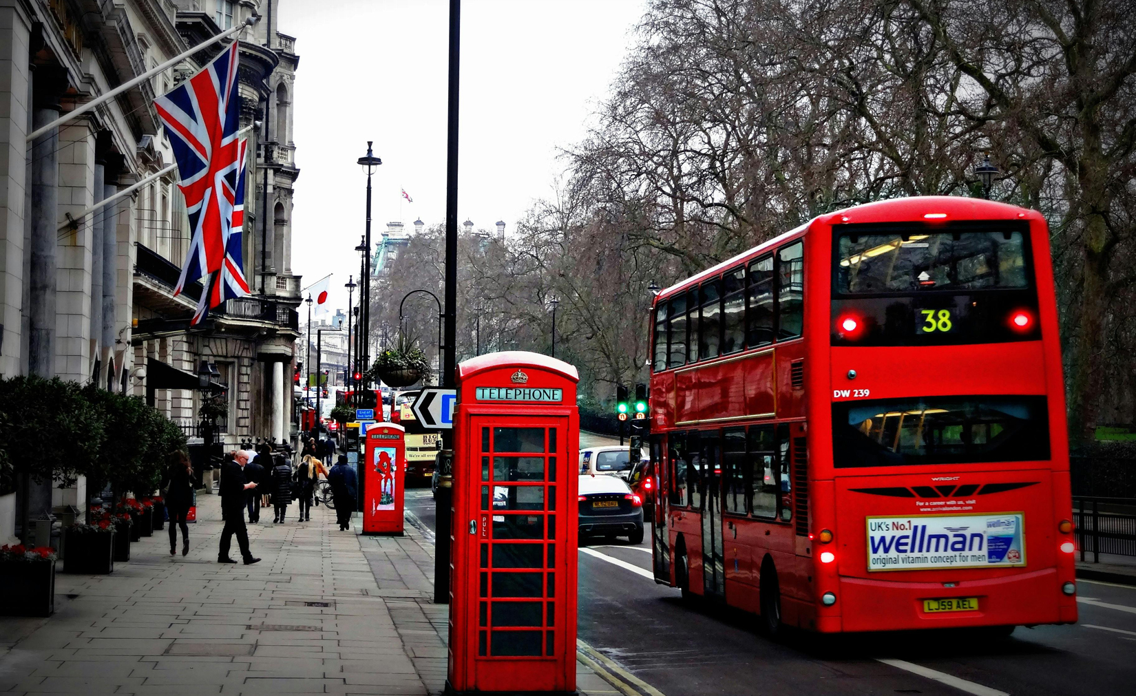 A vibrant urban scene featuring a red double-decker bus with route 38 passing by a classic red telephone booth on a busy London street. The backdrop includes historic buildings adorned with Union Jack flags, leafless trees, and pedestrians walking along the sidewalk. Image at Londonofficespace.com.