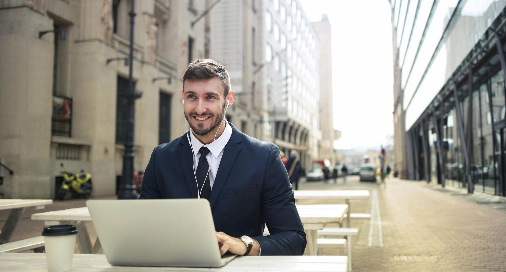 A man in a suit works on a laptop at an outdoor table in a bustling London street, with a mix of historic and modern buildings in the background. A coffee cup sits on the table, and he wears earphones while typing. The scene captures the essence of setting up an office in London. Image at Londonofficespace.com.