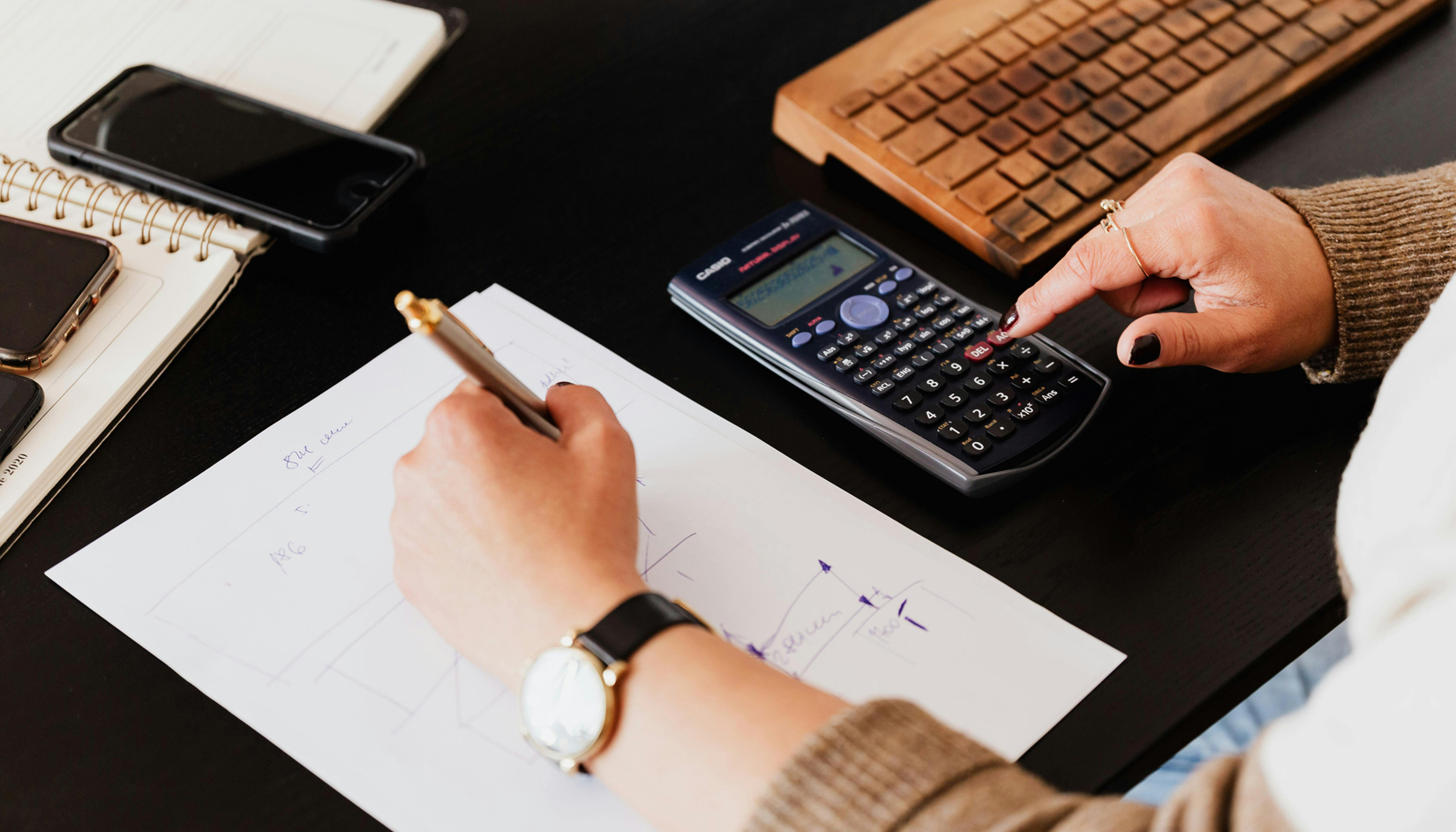 A person in a brown sweater works at a desk, writing on a sheet of paper with a pen and using a calculator, surrounded by a notebook, smartphones, and a wooden keyboard. The scene reflects the process of setting up an office in London, capturing the essence of financial planning. Image at Londonofficespace.com.